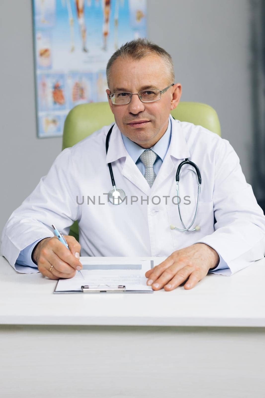 Portrait of confident old mature male head doctor physician in white medical uniform in glasses sitting at workplace. General practitioner looking at camera, posing in office by uflypro