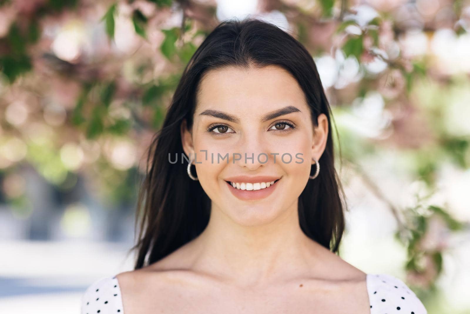 Portrait of a Gorgeous Dark Haired Woman Smiling Wearing Sammer Dress Charmingly while Standing at park on a background of sakura trees. Happy Young Woman Enjoys Life