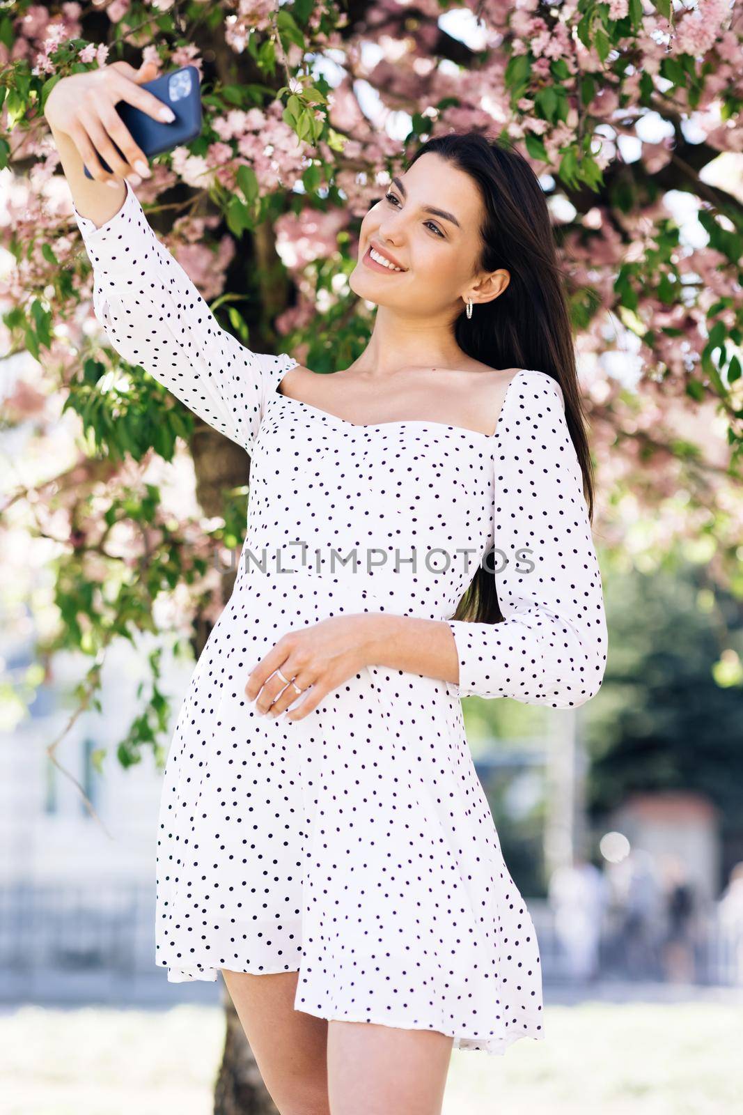 Vertical format of beautiful joyful young Caucasian woman in white dress posing to smartphone camera and taking selfie photo. Pretty stylish female tourist making photos with mobile phone by uflypro