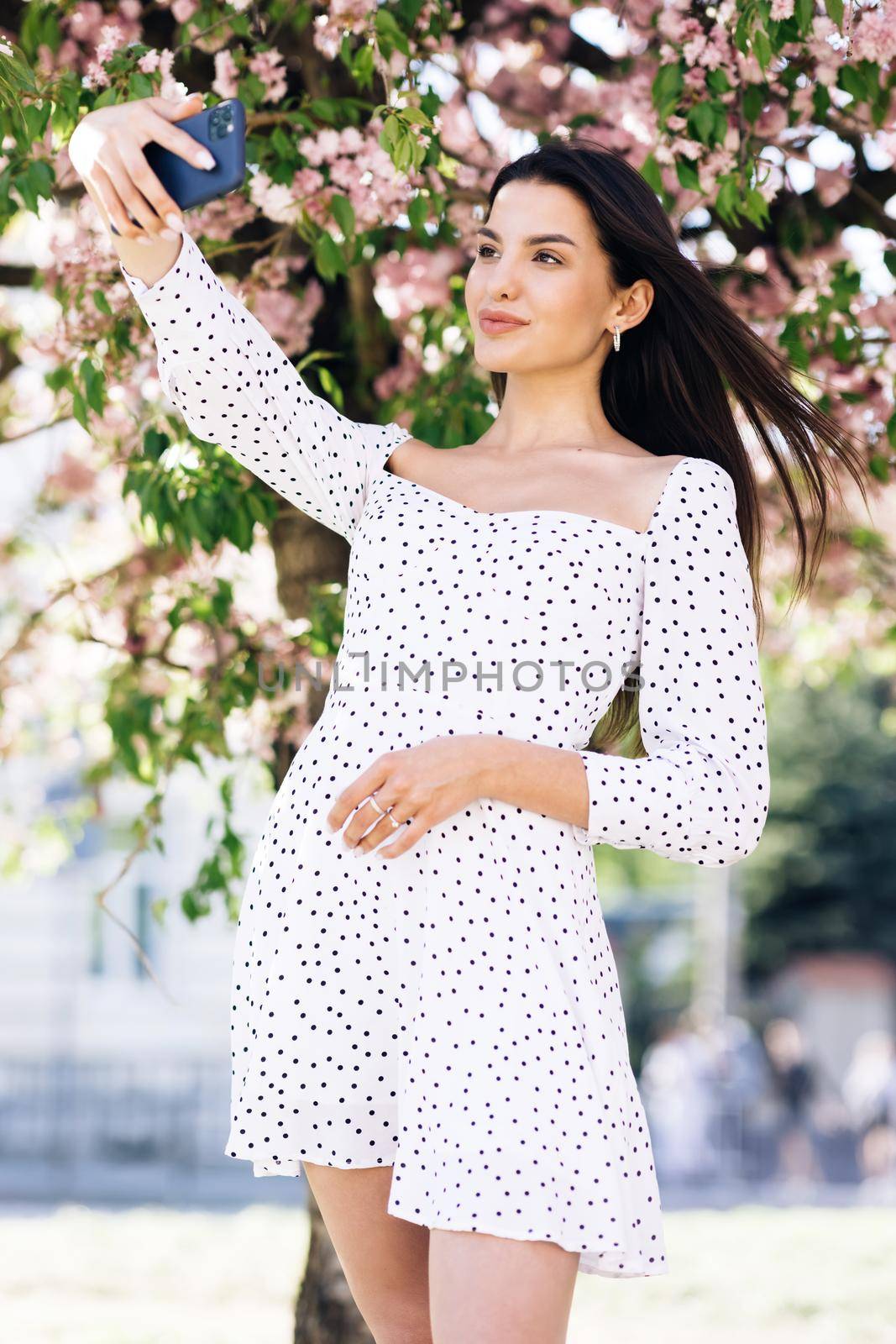 Female showing positive face emotions. Young woman in summer white dress taking selfie self portrait photos on smartphone. Model posing on park sakura trees background.