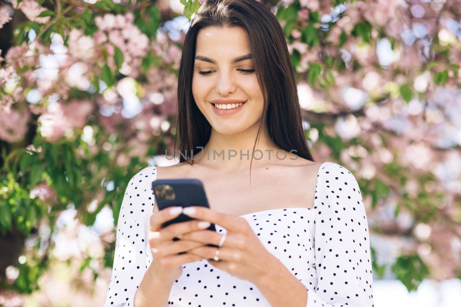 Smiling woman walks down the central park city street and uses her phone. Pretty summer woman in white dress walks down the street looking at her mobile phone by uflypro