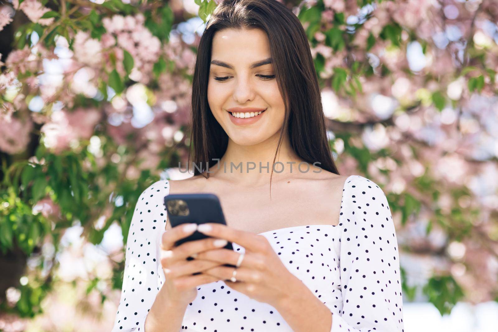 Smiling woman walks down the central park city street and uses her phone. Pretty summer woman in white dress walks down the street looking at her mobile phone.