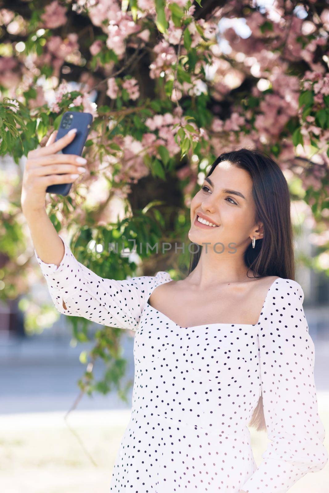 Smiling woman in summer white dress taking selfie self portrait photos on smartphone. Model posing on park sakura trees background. Female showing positive face emotions. by uflypro