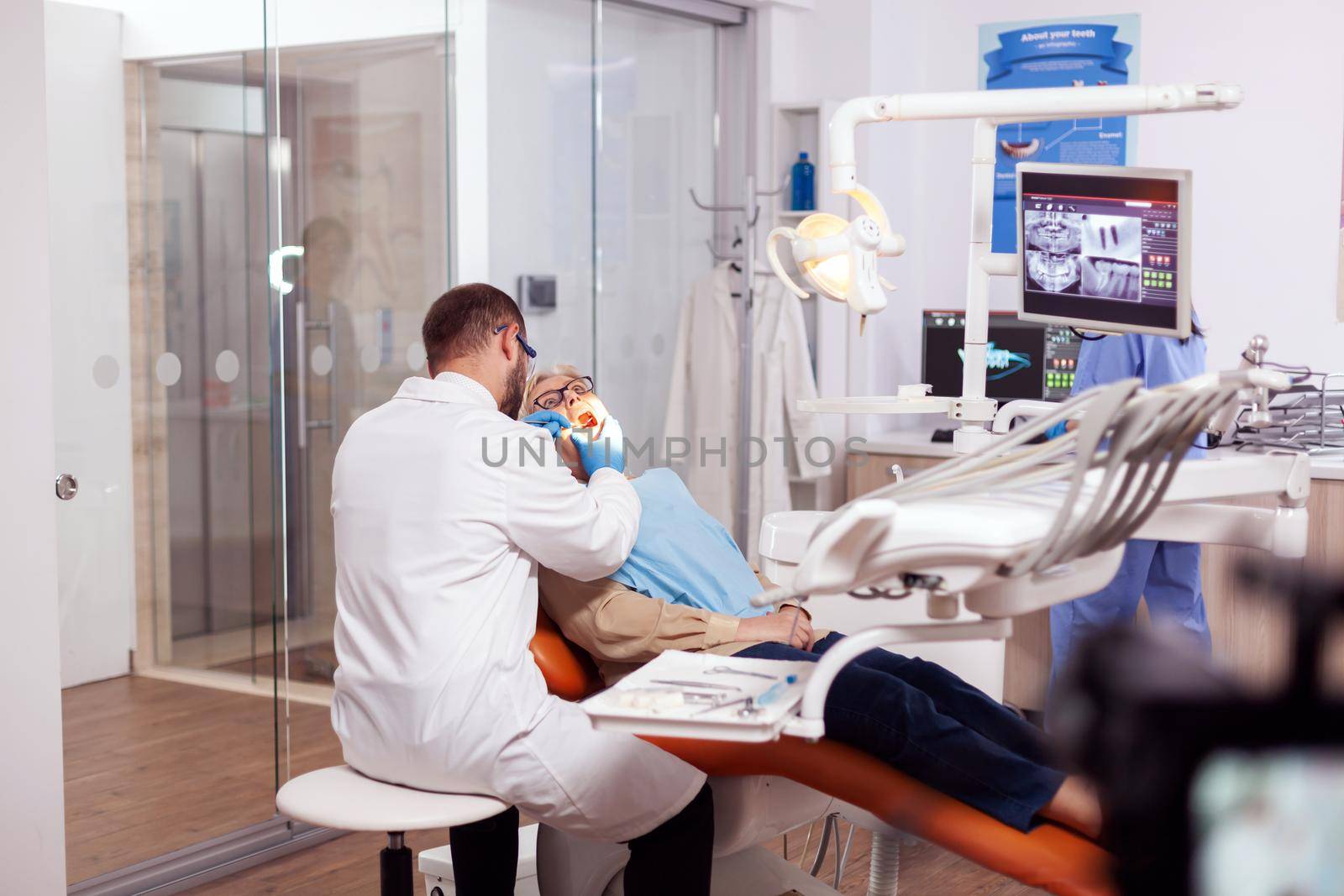 Dentist examining senior patient mouth using angled mirror and scaler during treatment. Elderly patient during medical examination with dentist in dental office with orange equipment.