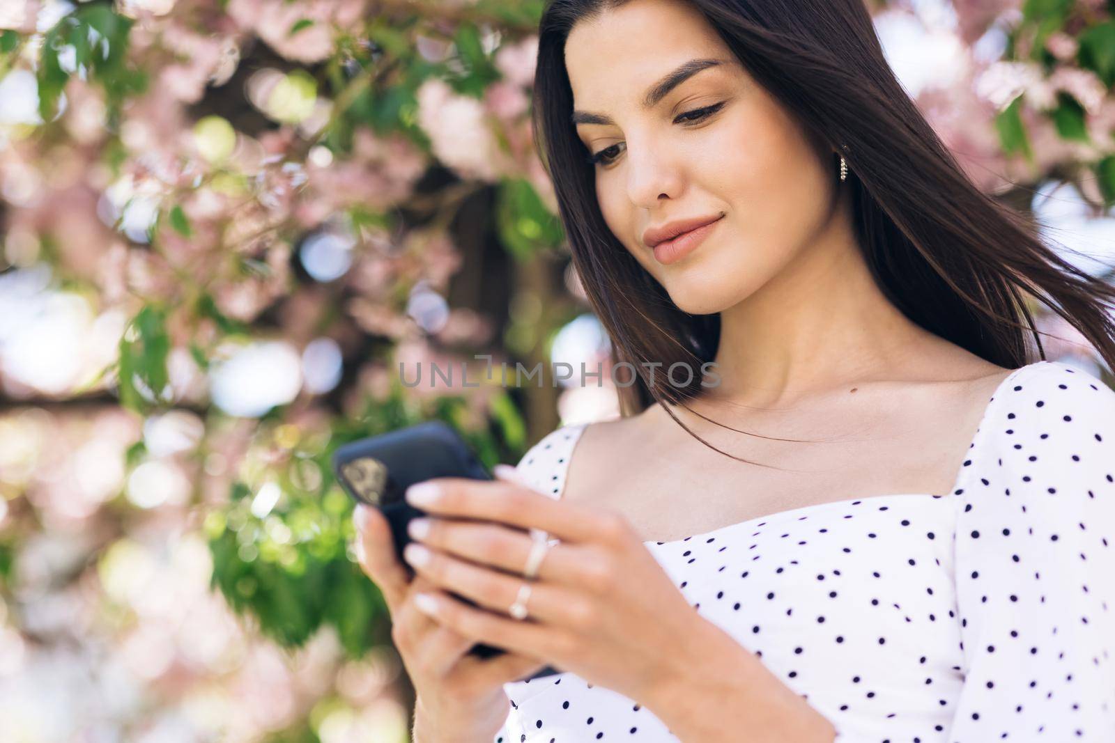 Cheerful woman looking at mobile phone in the street urban people with cellphone in hand. Pretty summer woman in white dress walks down the street looking at her mobile phone. by uflypro