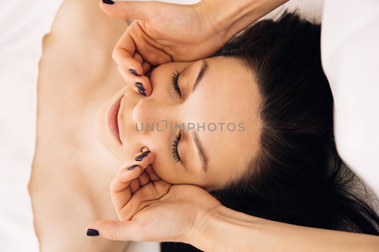 Cosmetologist making skin care procedure in beauty salon, portrait of girl closeup. Beautician doctor is applying facial cleansing foam on woman's face massaging skin in cosmetology clinic.