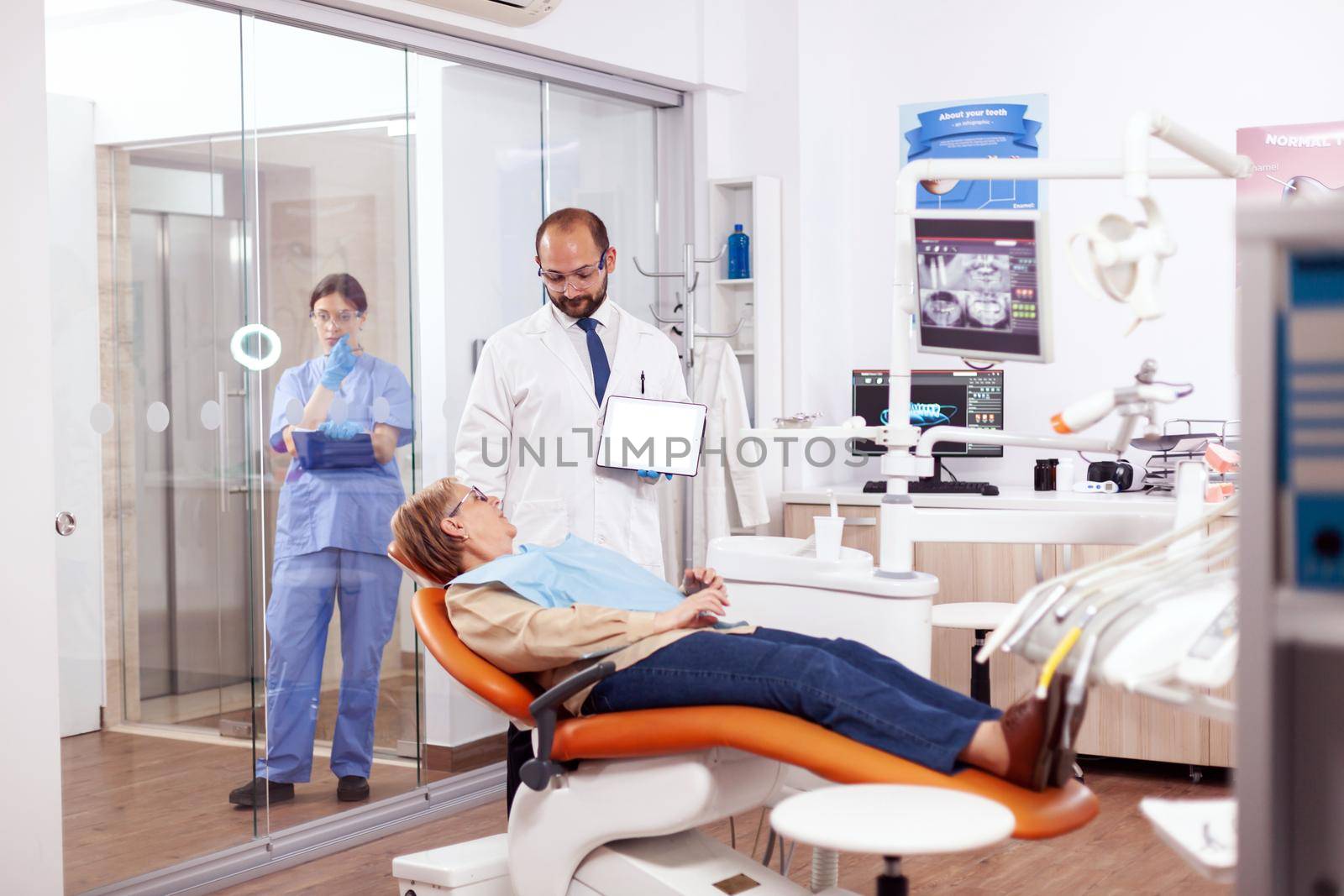 Dentist discussing with senior woman in cabinet holding tablet pc with copy sapce available. Medical teeth care using digital device with patient radiography on tablet pc near patient standing up.