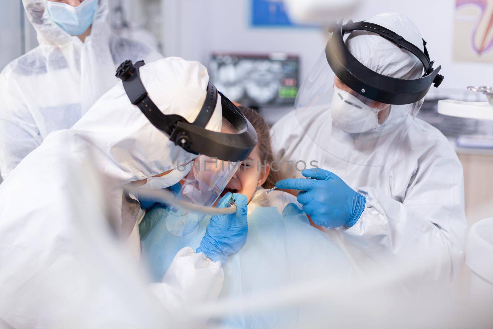 Caucasian little girl having dental treatment at dentist's office sitting on chair with bib. Stomatology team wearing ppe suit during covid19 doing procedure on child teeth.