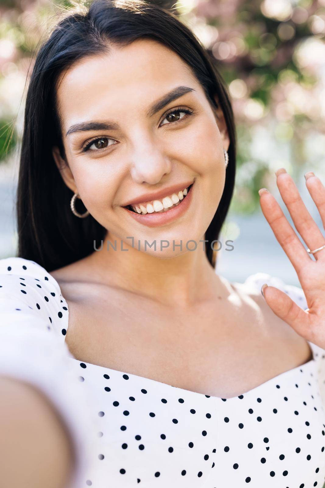 Vertical photo of smiling girl having video call on smartphone in city park, friendly woman waving hand at camera, pretty woman video chatting at phone, stylish blogger making video on phone outdoor.