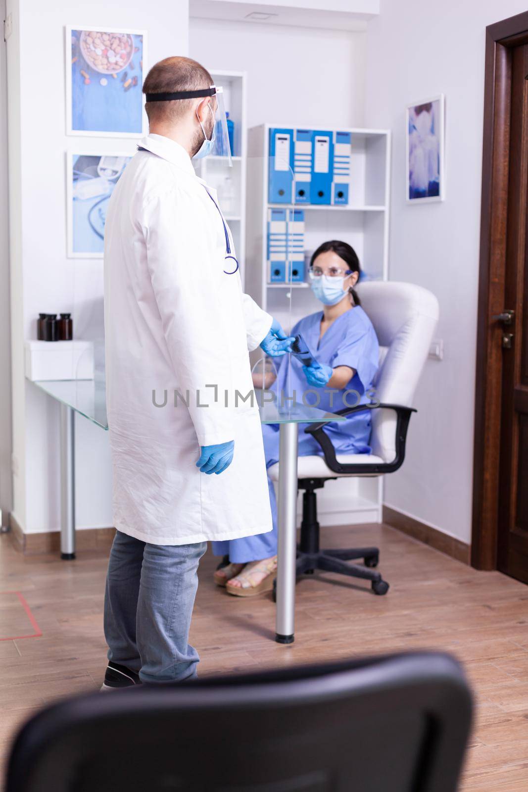Nurse giving doctor radiography of invalid patient and wearing face mask in hospital waiting room. Assistant working on reception computer.