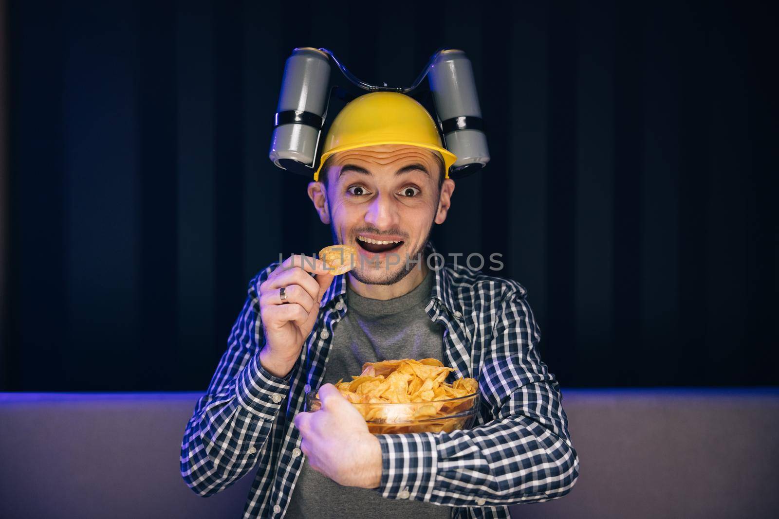 Caucasian man rests at home and watches TV shows or sports news on TV screen. Man with beer helmet on the head eats chips while sitting at home on couch, in evening he watches movies at home on TV.