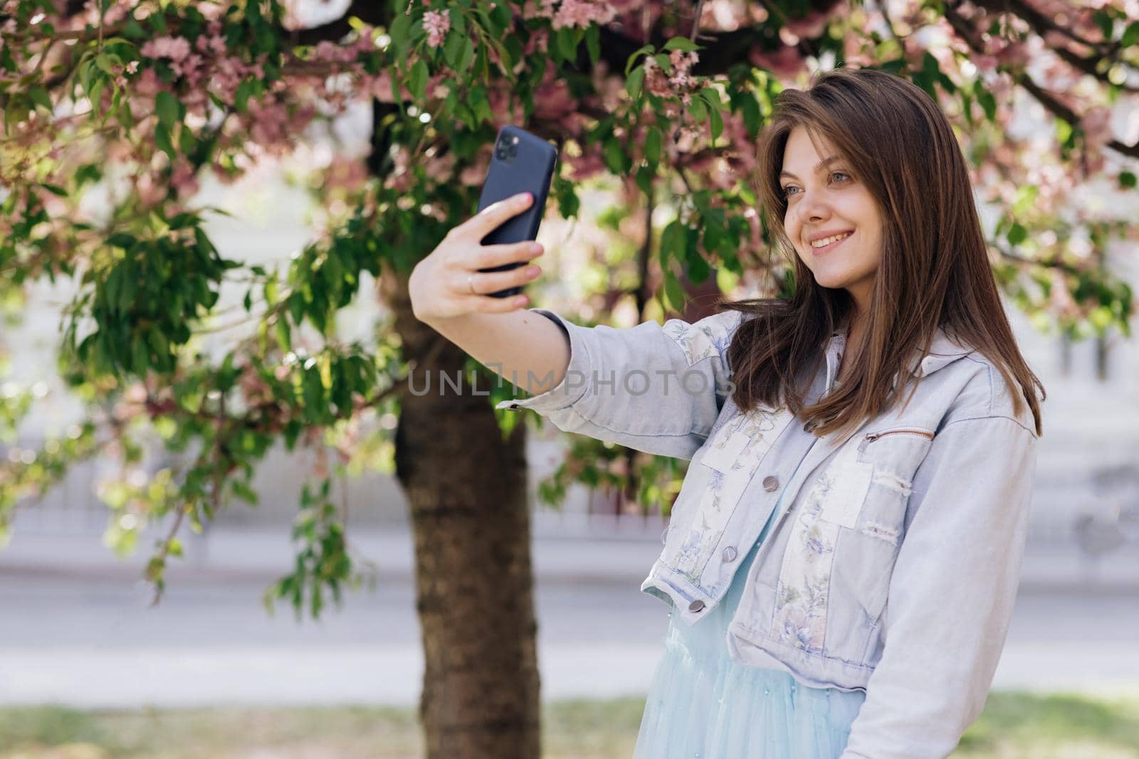 Young smiling woman taking selfie self portrait photos on smartphone. Model posing on park sakura trees background. Female showing positive face emotions. by uflypro