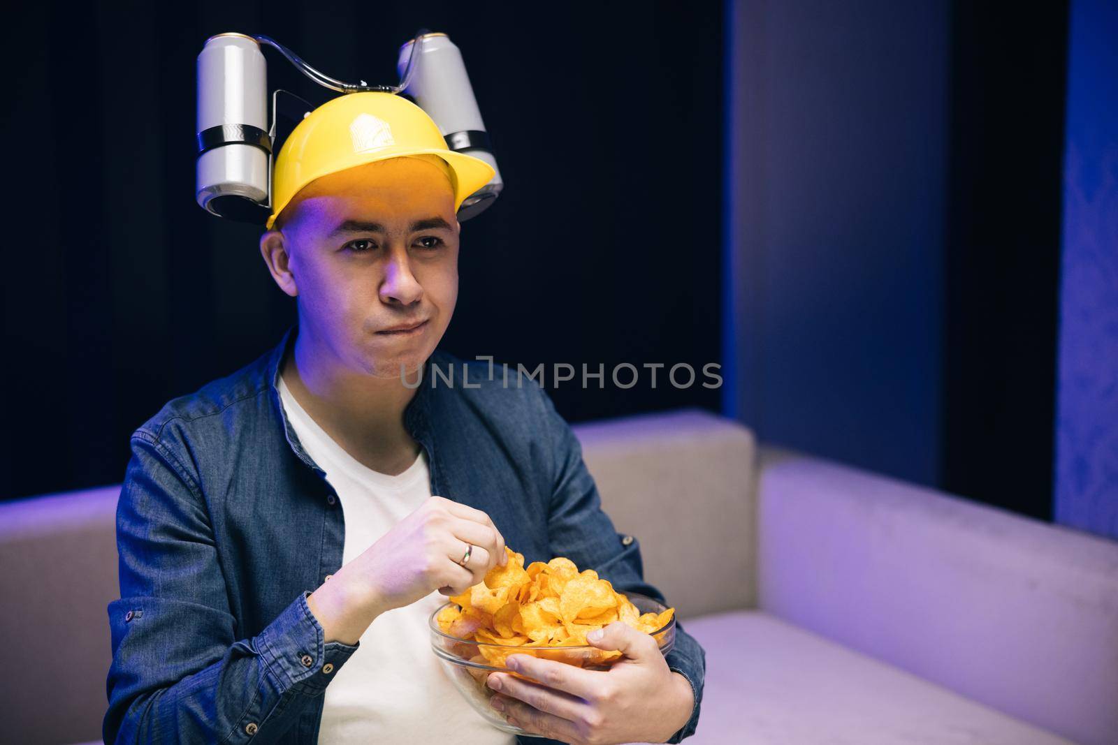 Businessman with beer helmet on the head relaxing watching a comedy while eating chips and drinking beer sitting on couch.