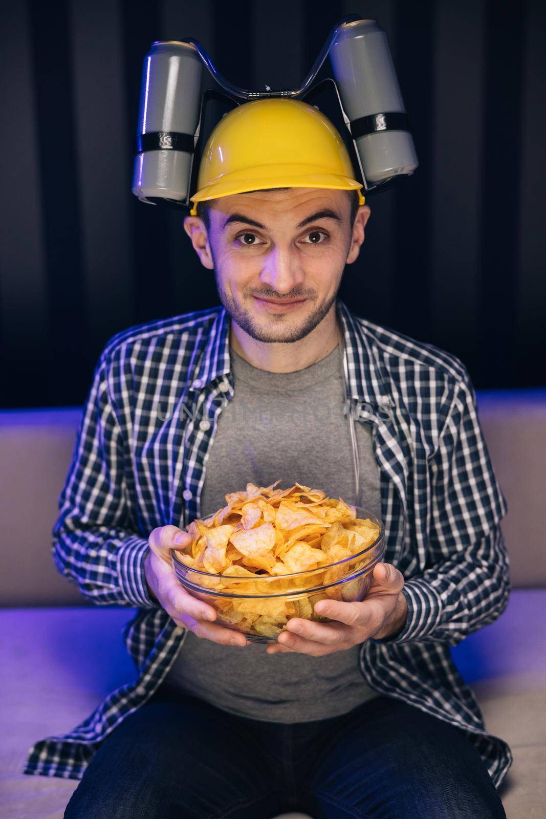 Handsome man with beer helmet on the head watching TV and eating potato chips on the couch at home. Young male sits on a sofa and eats snacks while watching a movie by uflypro