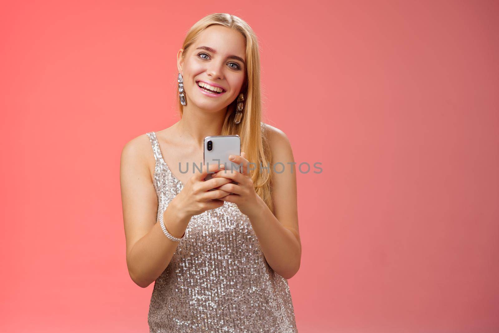 Lifestyle. Charming elegant young 25s woman in silver dress brilliant accessorize holding smartphone messaging using app order taxi after nightclub friends party standing carefree smiling camera joyfully.