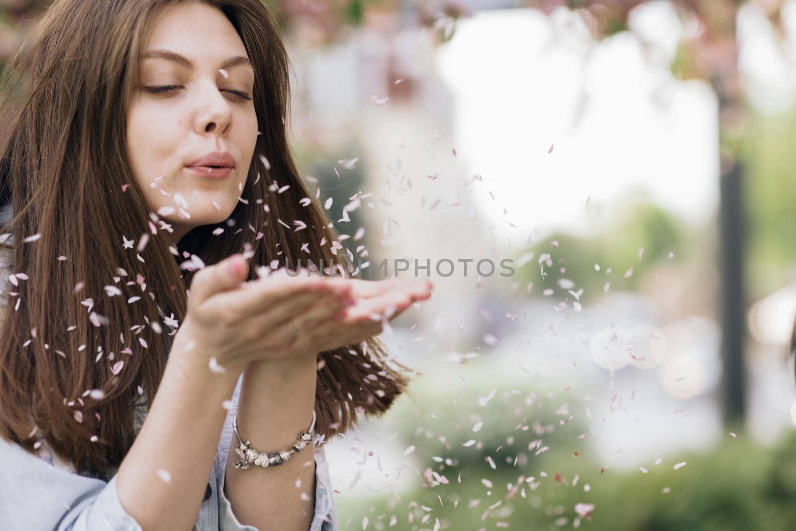 Woman Enjoying Spring Blossom. Happy Smiling Girl blowing sakura flower petals, making them fly to camera. Sunny Spring Outdoors Activity. Spring blossom of sakura tree in city park.