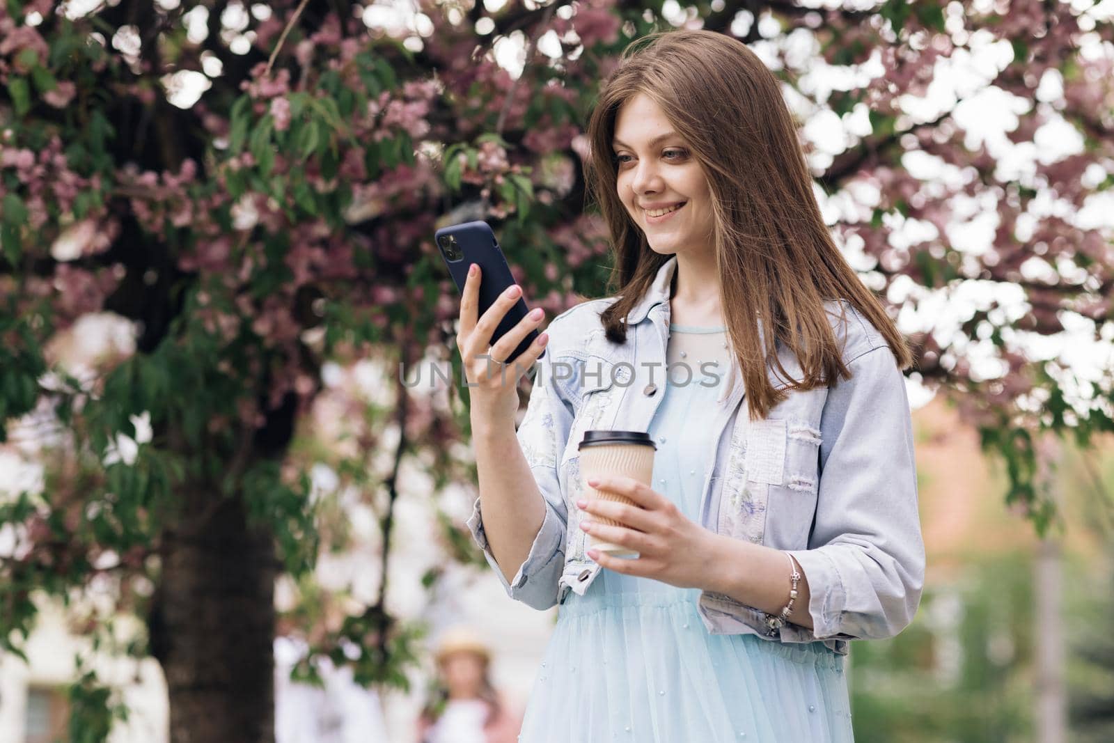 Woman with charming Smile walking in the Park and Using Mobile Phone Wearing Stylish Outfit. Girl Using Smartphone and Drinking Coffee. Communication, online shopping, social network concept by uflypro