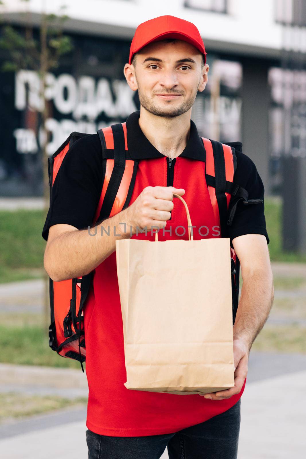 Outdoor portrait of delivery man with red uniform holding food bags waiting for customer. Happy young courier is proud of his job smiling standing in the street. Home delivery.