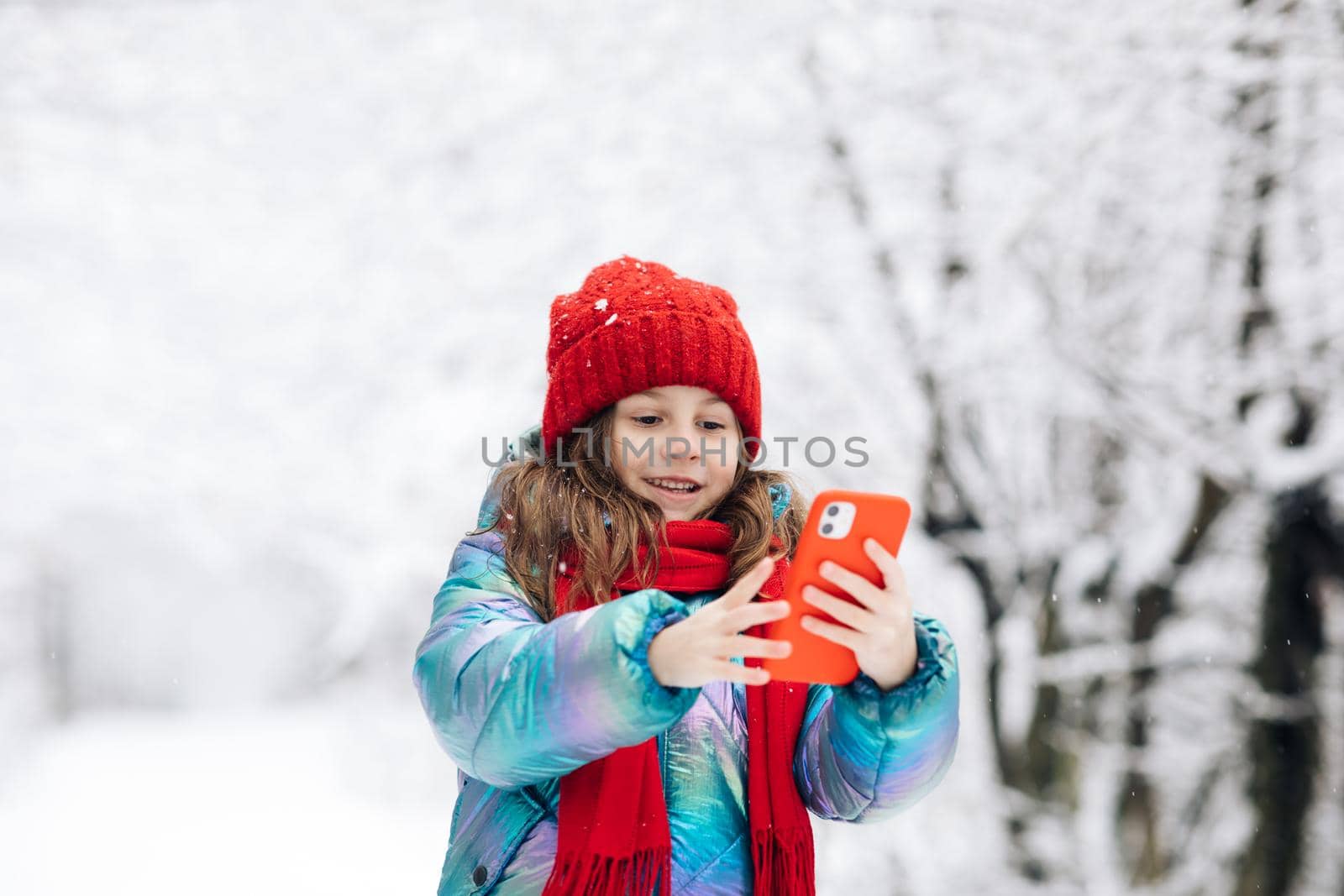 Cute little girl taking a selfie in the winter forest. Winter travel with children concept. Adorable happy smiling kid girl enjoy making selfie camera shot by smartphone.