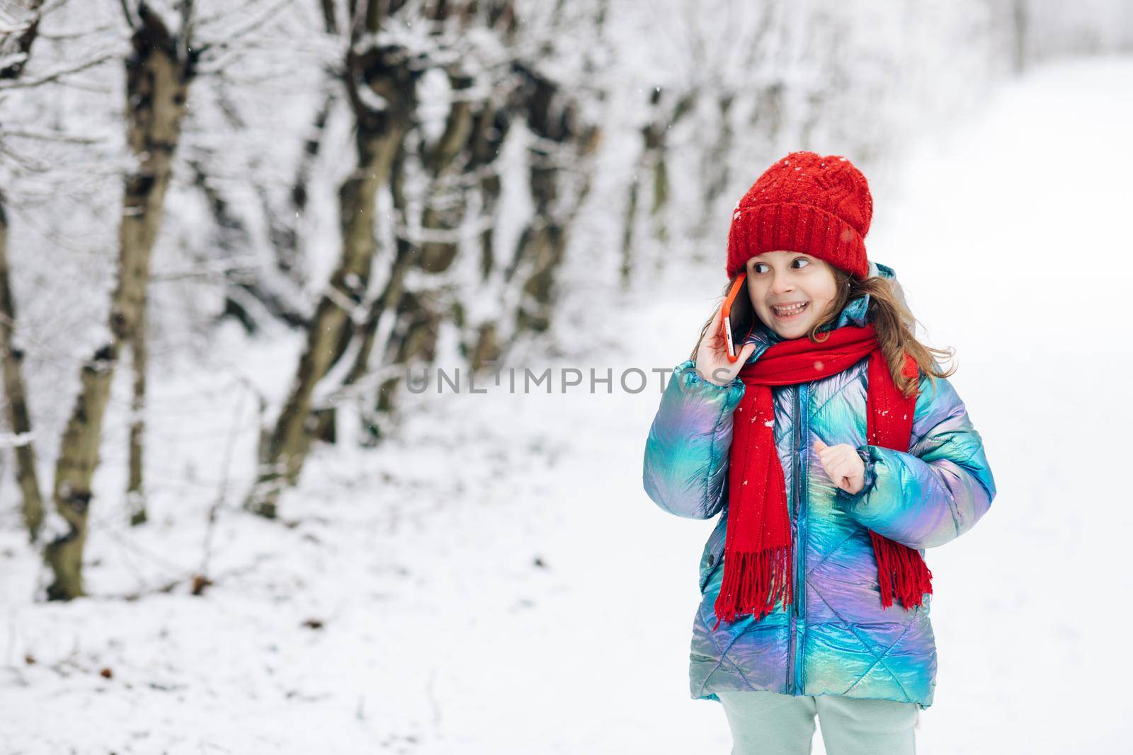 Young beautiful child girl in winter hat talking emotionally and surprised talking on cell phone. Young curly girl talking on her mobile phone while walking in the winter city street.