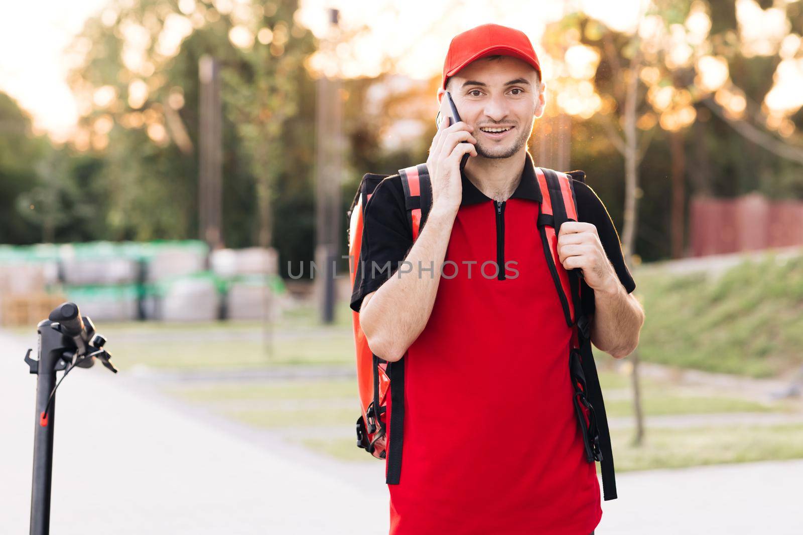 Male courier with isothermal food case box arrives to the entrance to the house and calls for client. Food delivery guy with red backpack deliver orders.