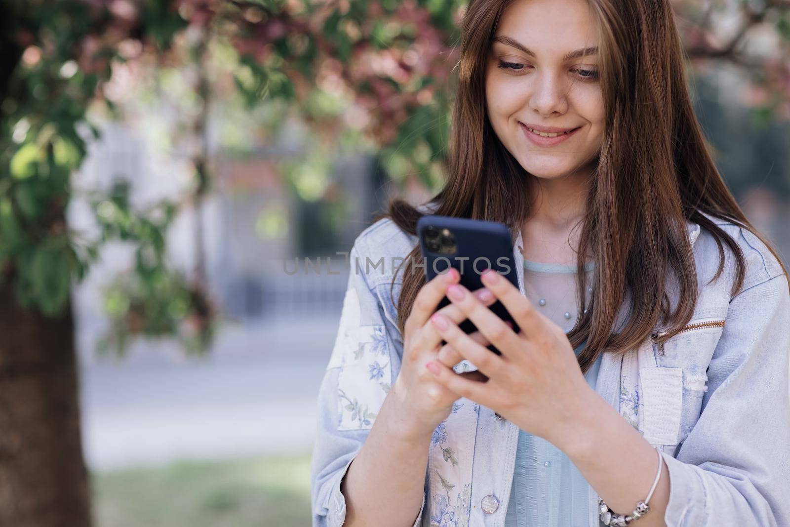 Smiling woman walks down the central park city street and uses her phone. Pretty summer woman in white dress walks down the street looking at her mobile phone by uflypro