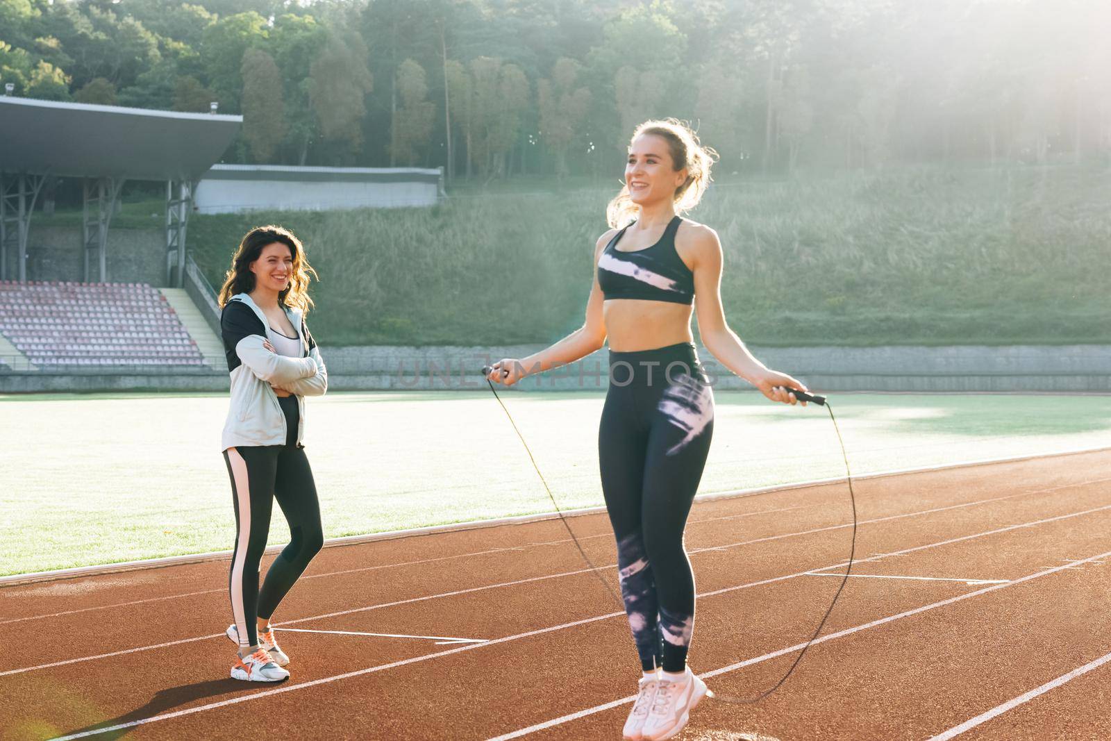 Sporty female with a good figure jumps rope on sports track of stadium. Athletic woman with personal instructor skipping rope as part of her fitness workout. Exercising strength cardio and power by uflypro