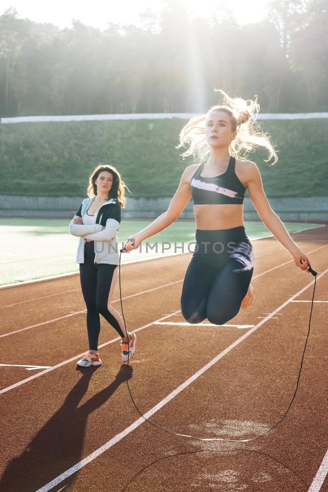 Athletic woman with personal instructor skipping rope as part of her fitness workout. Sporty female with a good figure jumps rope on sports track of stadium. Exercising strength cardio and power by uflypro