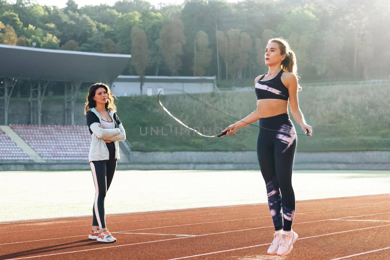 Athletic caucasian woman with personal trainer jumping rope as part of her fitness workout. Sporty female with a good figure jumps rope on sports track of stadium. Exercising strength cardio and power