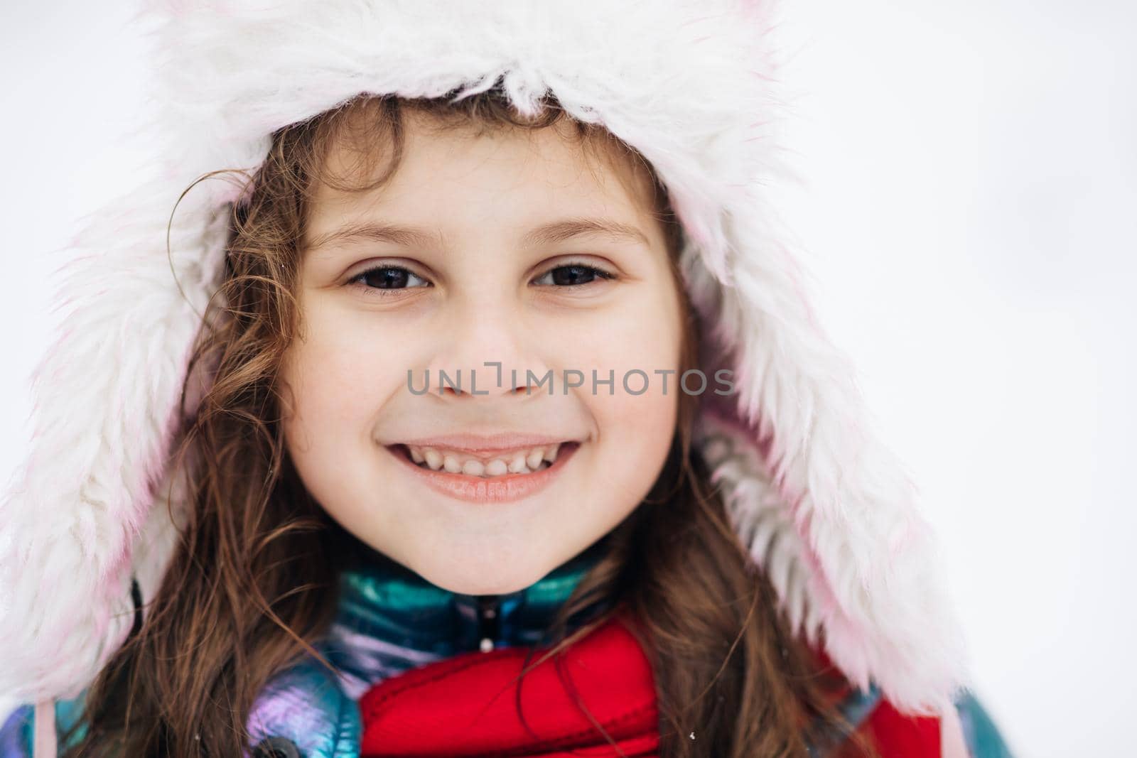 Funny cute girl laughing outdoors in winter day. Pretty little girl smiling with long hair in red hat and scarf standing in the white snowy park or forest at winter time