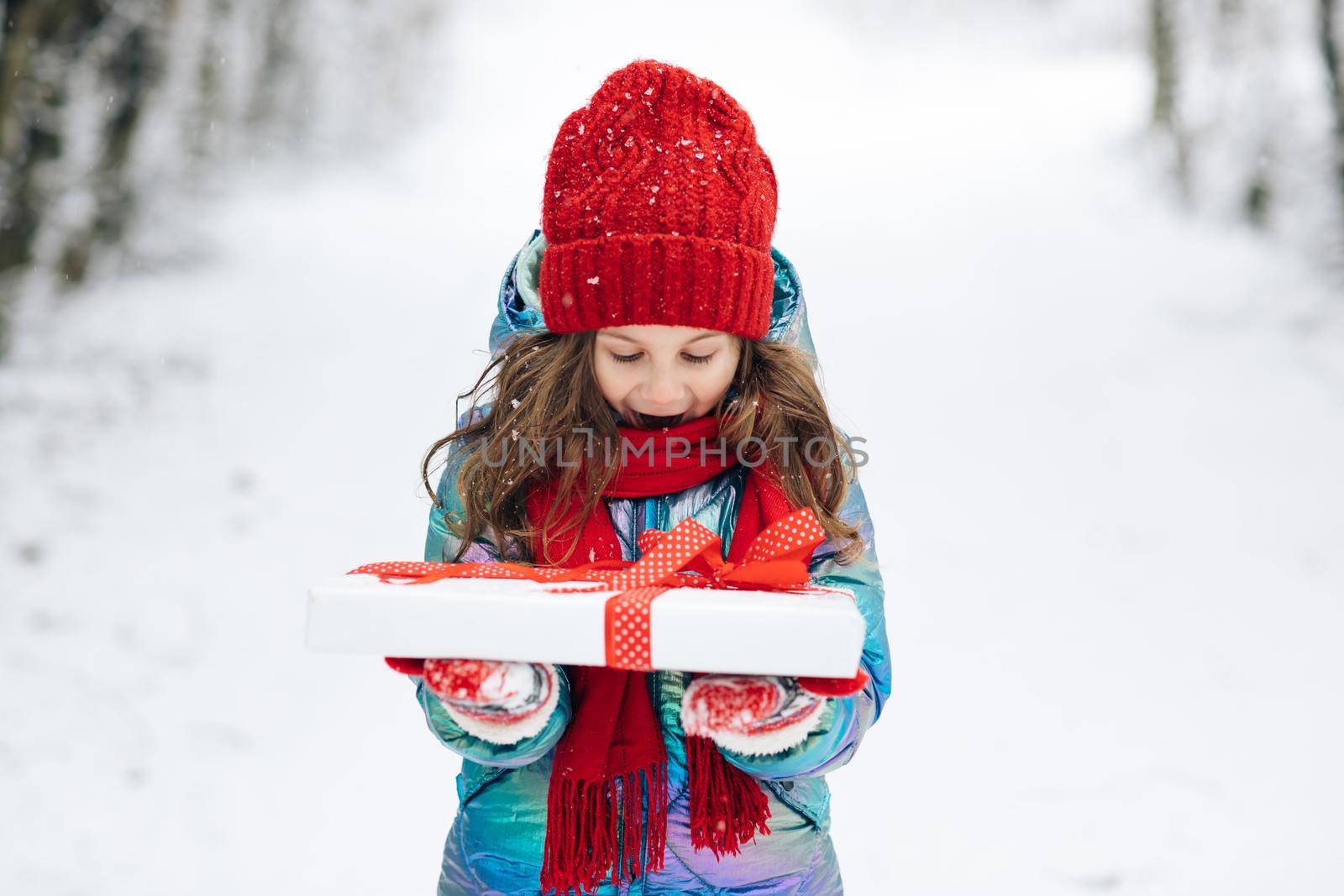 Child holding gift box and surprise face. Child in red hat with Christmas gift box in snow. Winter outdoor fun. Kid play in snowy park. by uflypro