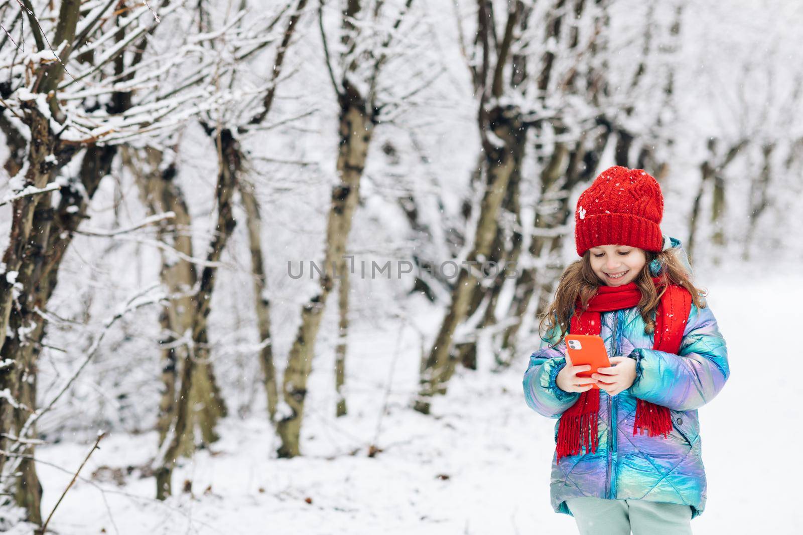 Little girl rolling up a funny video in smartphone. Happy child playing at winter forest use a smartphone look at the screen of a cell phone, watch cartoons.