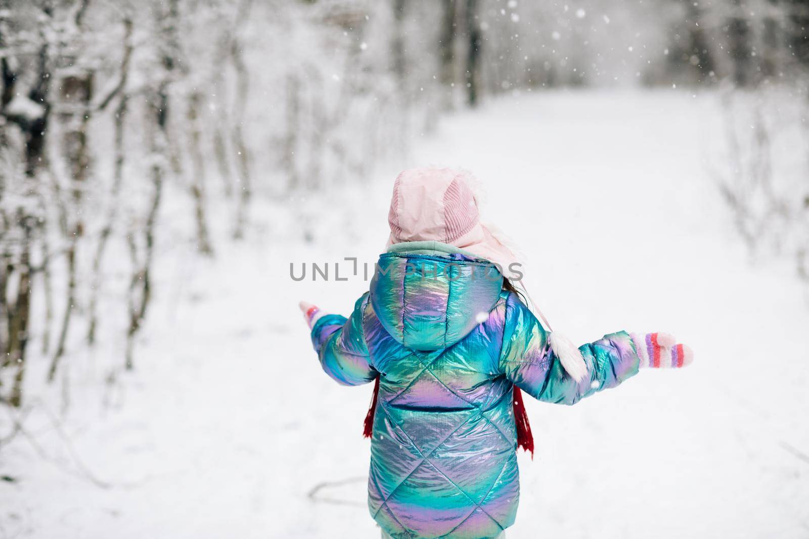 Back view happy laughing child, cute girl in a colorful clothing and hat, running in a snowy winter park catching snowflakes. Little winter girl in fairy ice forest. Walk in winter forest.