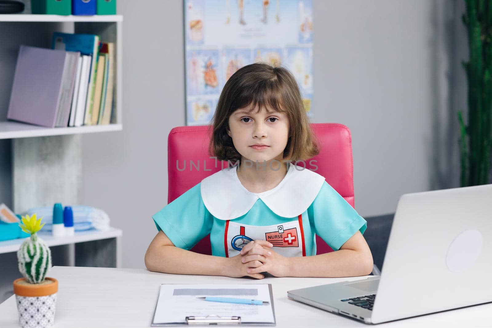 Funny little girl looking at camera are sitting on the couch at home. Cute kid child with pretty face. Concept of a happy childhood. Portrait of girl in medic uniform looking at camera