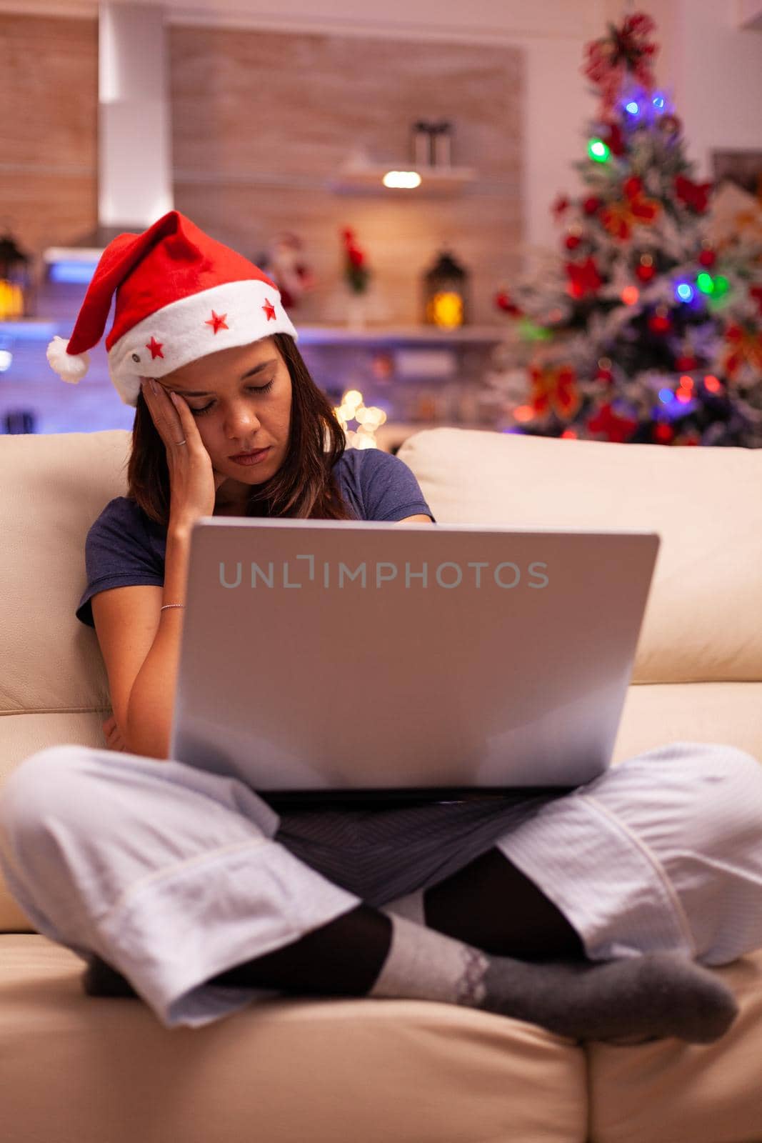 Tired exhausted woman falling asleep on couch in xmas decorated kitchen while reading business email on computer. Caucasian female celebrating christmas holiday enjoying winter season