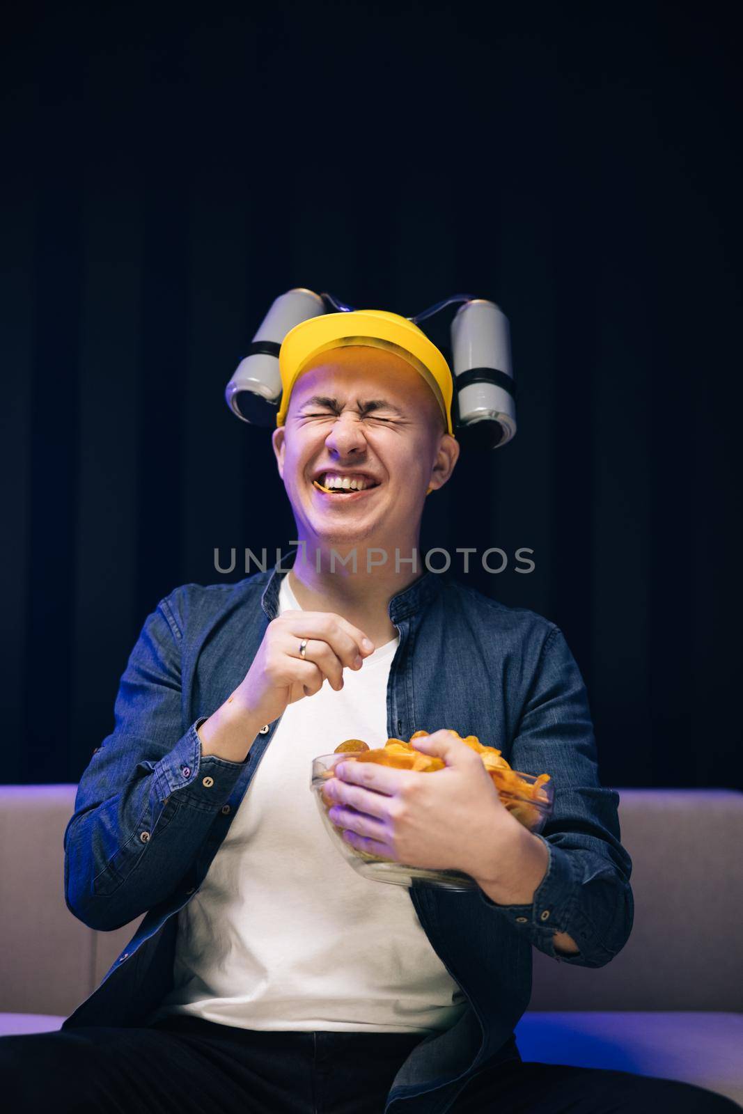 Young male sits on a sofa and eats snacks while watching a movie. Handsome man with beer helmet on the head watching TV and eating potato chips on the couch at home by uflypro