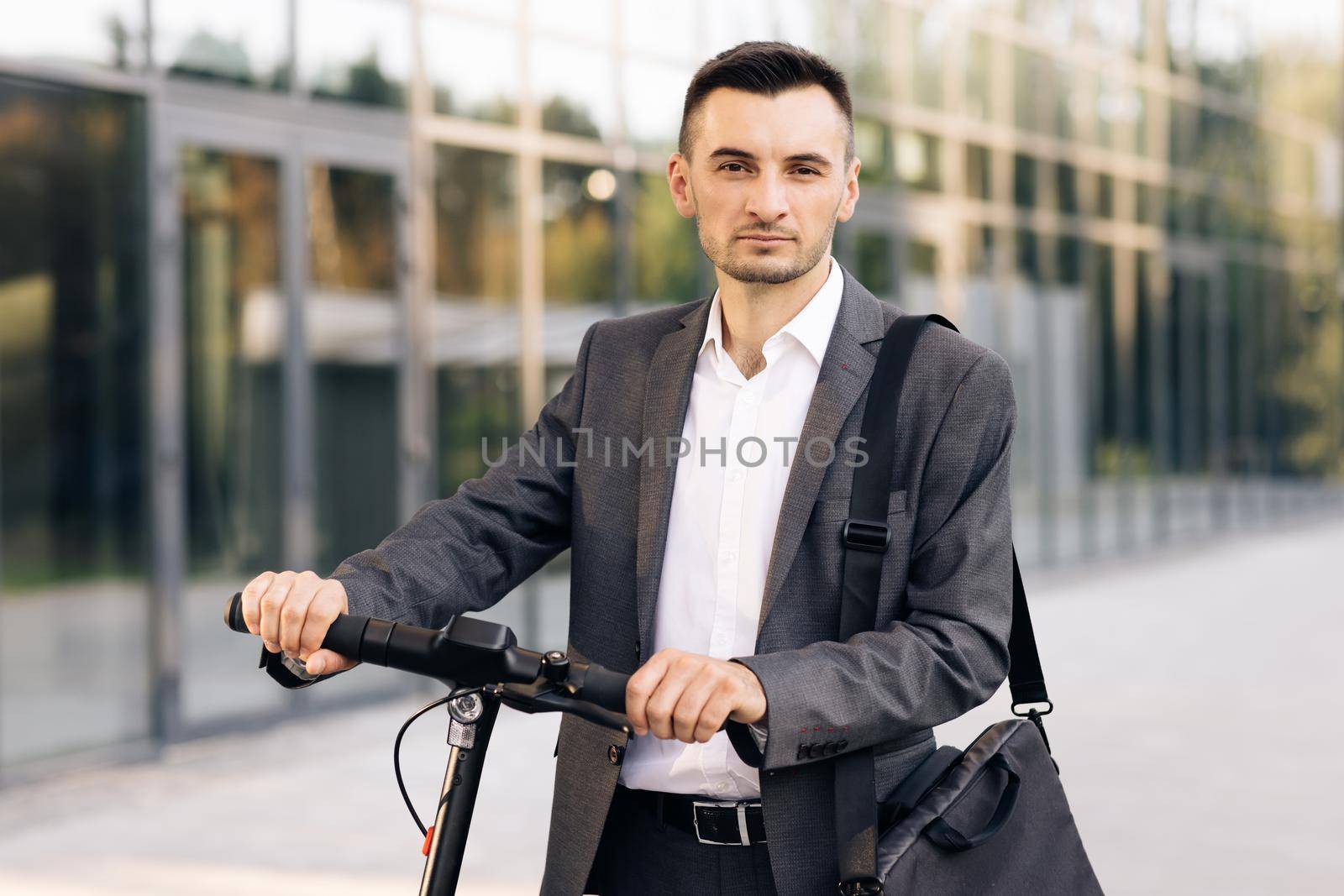 Stylysh man on vehicle outdoors. Modern urban alternative transport. Portrait of confident businessman standing with electric scooter and looking at camera. E-Scooter rider rent personal eco transport.