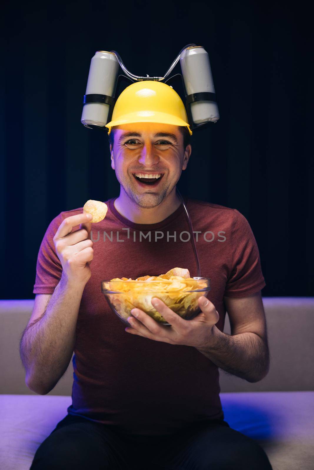 Handsome man with beer helmet on the head watching TV and eating potato chips on the couch at home. Young male sits on a sofa and eats snacks while watching a movie.
