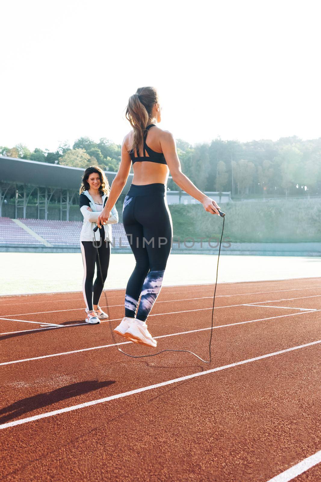 Woman with personal trainer jumping rope as part of her fitness workout. Sporty female with a good figure jumps rope on sports track of stadium. Exercising strength cardio and power.