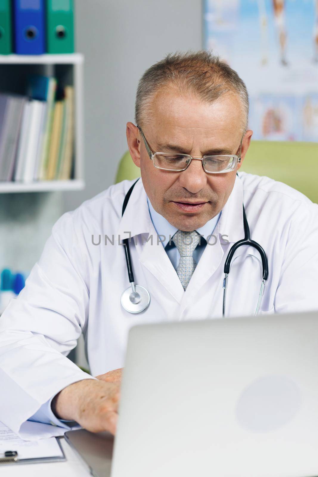 Calm Family Medical Doctor in Glasses is Working on a Laptop Computer in a Health Clinic. Physician in White Lab Coat is Browsing Medical History Behind a Desk in Hospital Office by uflypro