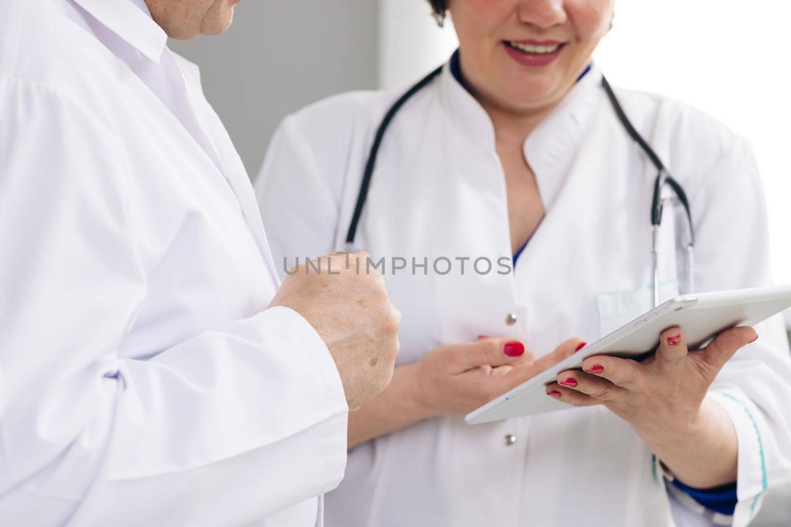 Caucasian person and his female colleagues use electronic device and work together while standing indoors. Closeup view of man and woman doctors using tablet during working day at clinic by uflypro
