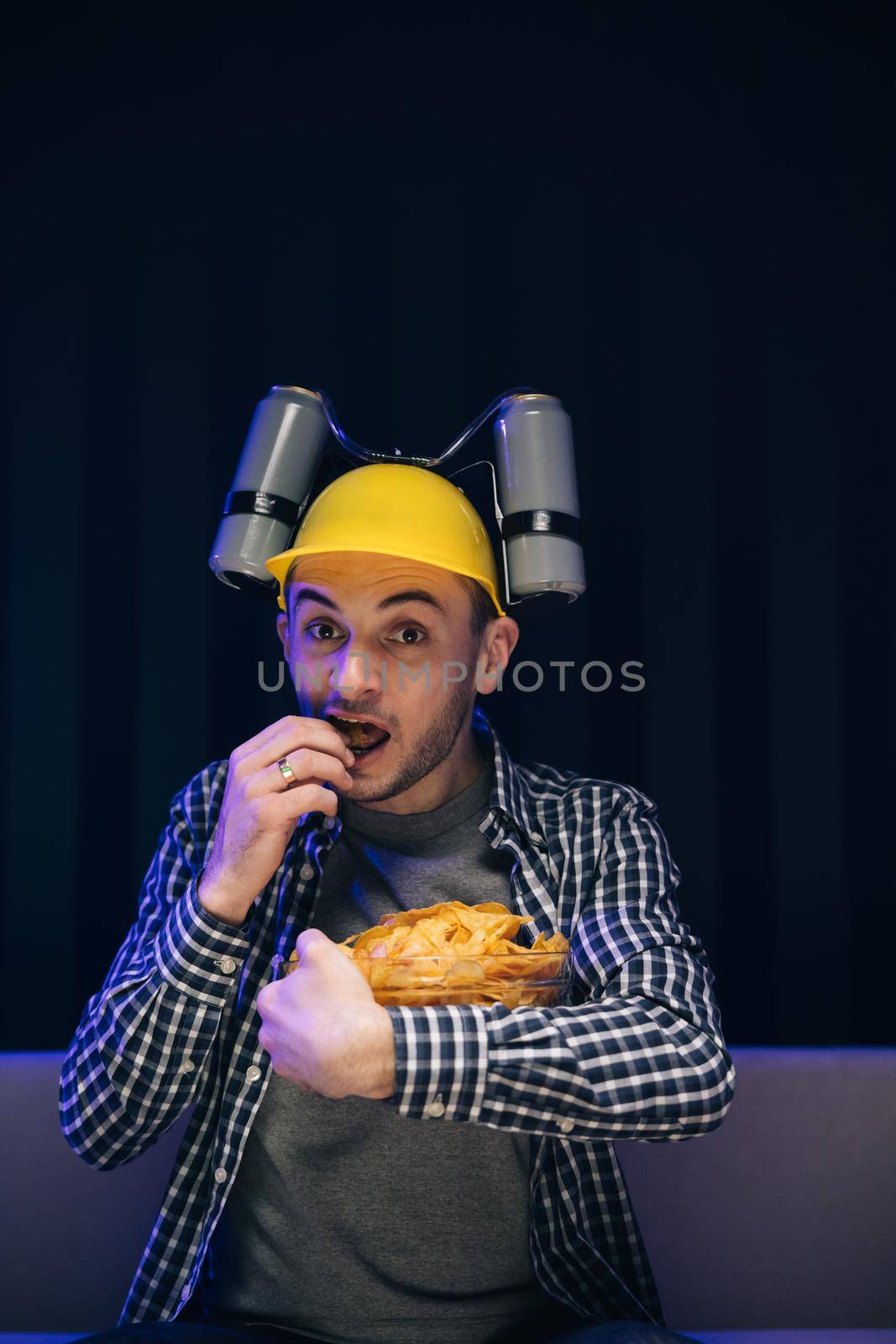 Close up of the young funny man with beer helmet on the head eating chips while watching movie on the sofa in the living room.