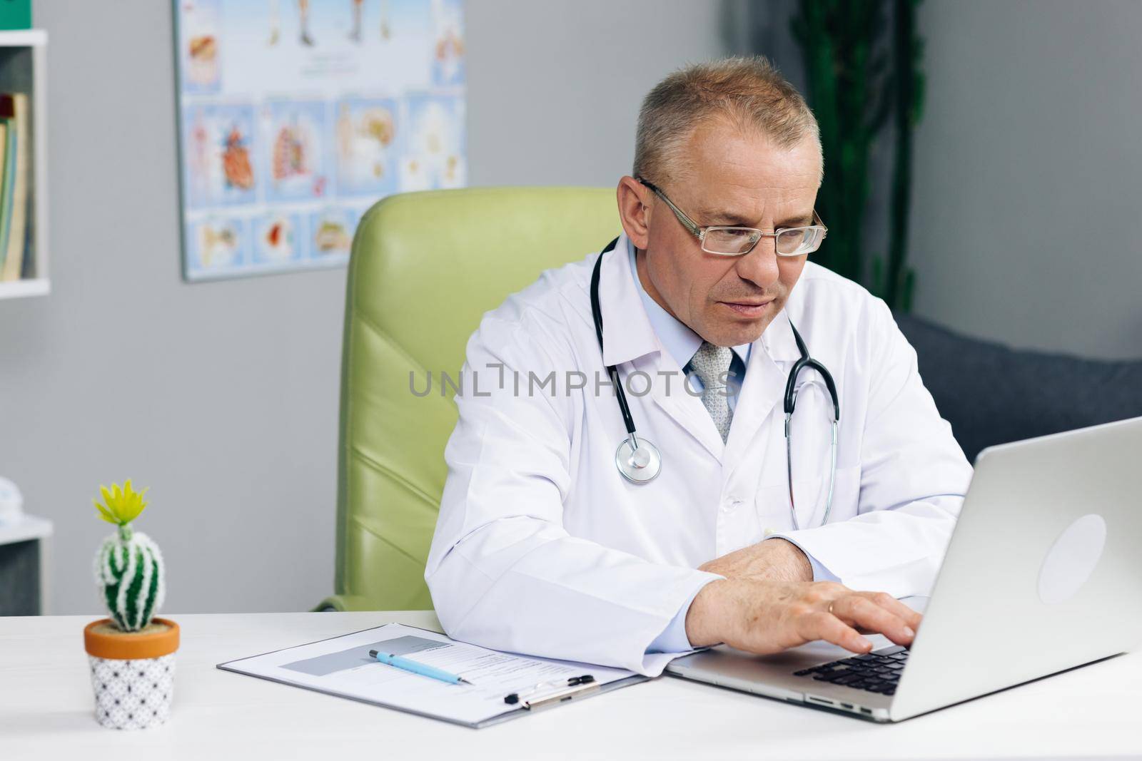 Physician in White Lab Coat is Browsing Medical History Behind a Desk in Hospital Office. Calm Family Medical Doctor in Glasses is Working on a Laptop Computer in a Health Clinic.