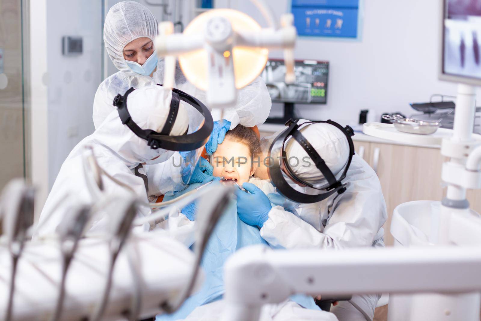 Little girl screaming in the course of painfull cavity treatment at dentist office dressed in protective suit. Stomatology team wearing ppe suit during covid19 doing procedure on child teeth.