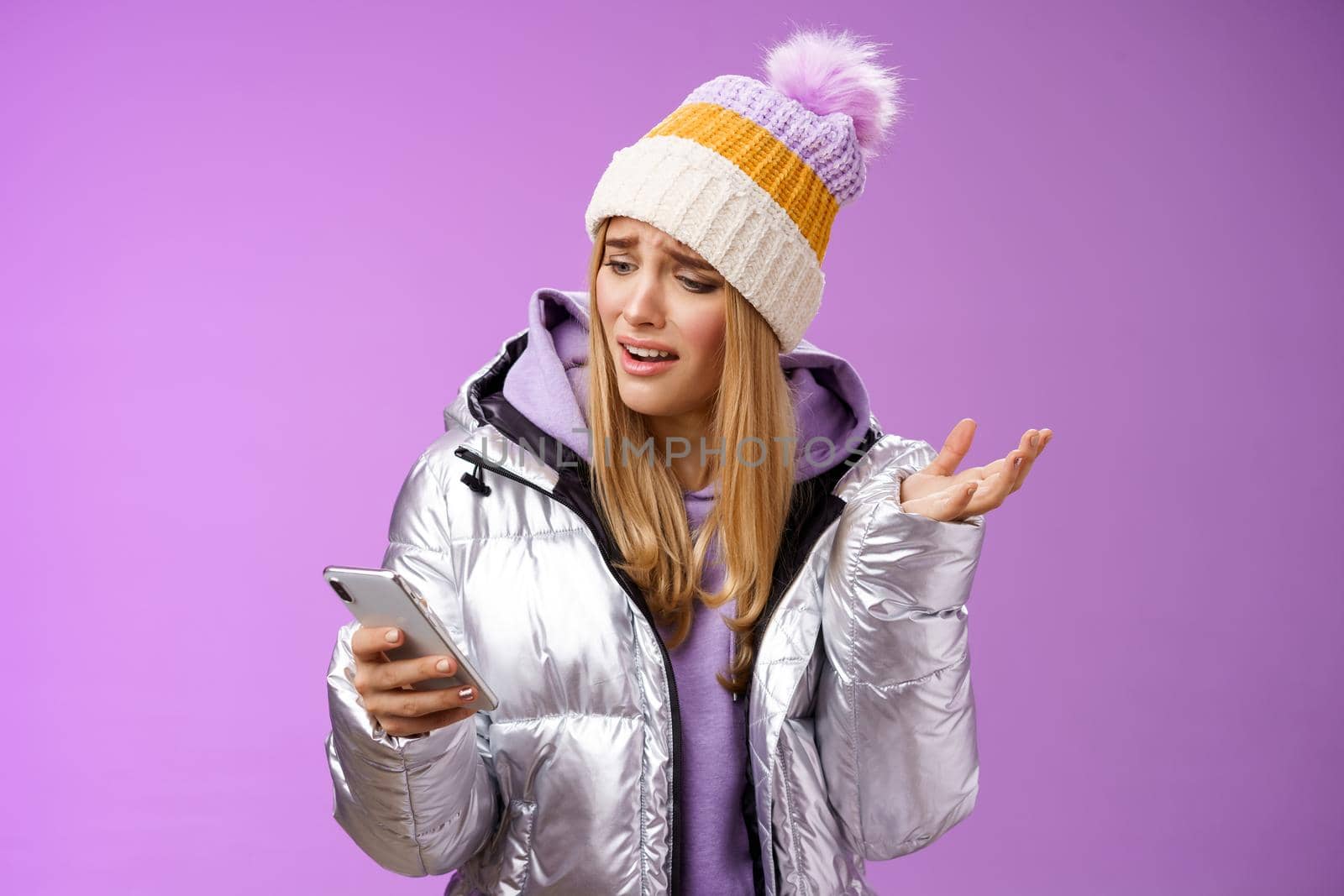 Upset disappointed attractive whining gloomy blond girl in silver jacket standing outside hat holding smartphone shrugging raising hand dismay complaining slow mobile internet, purple background by Benzoix