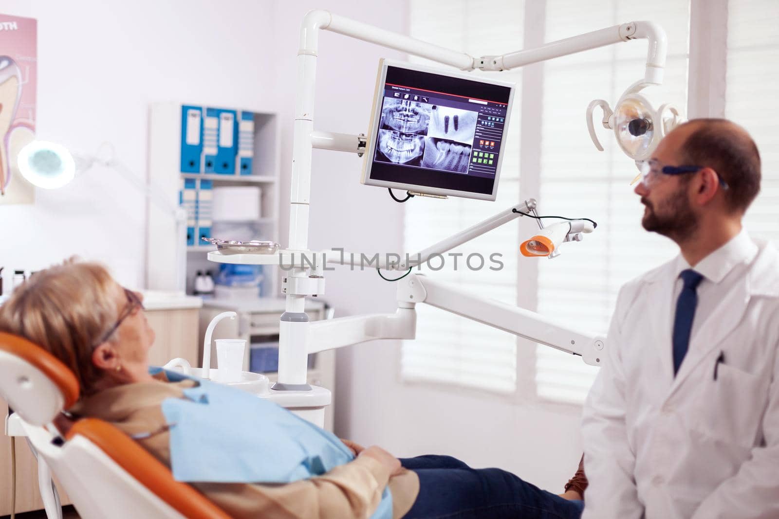 Stomatolog talking about teeh treatment to senior woman in dentist cabient. Medical teeth care taker pointing at patient radiography on screen sitting on chair.