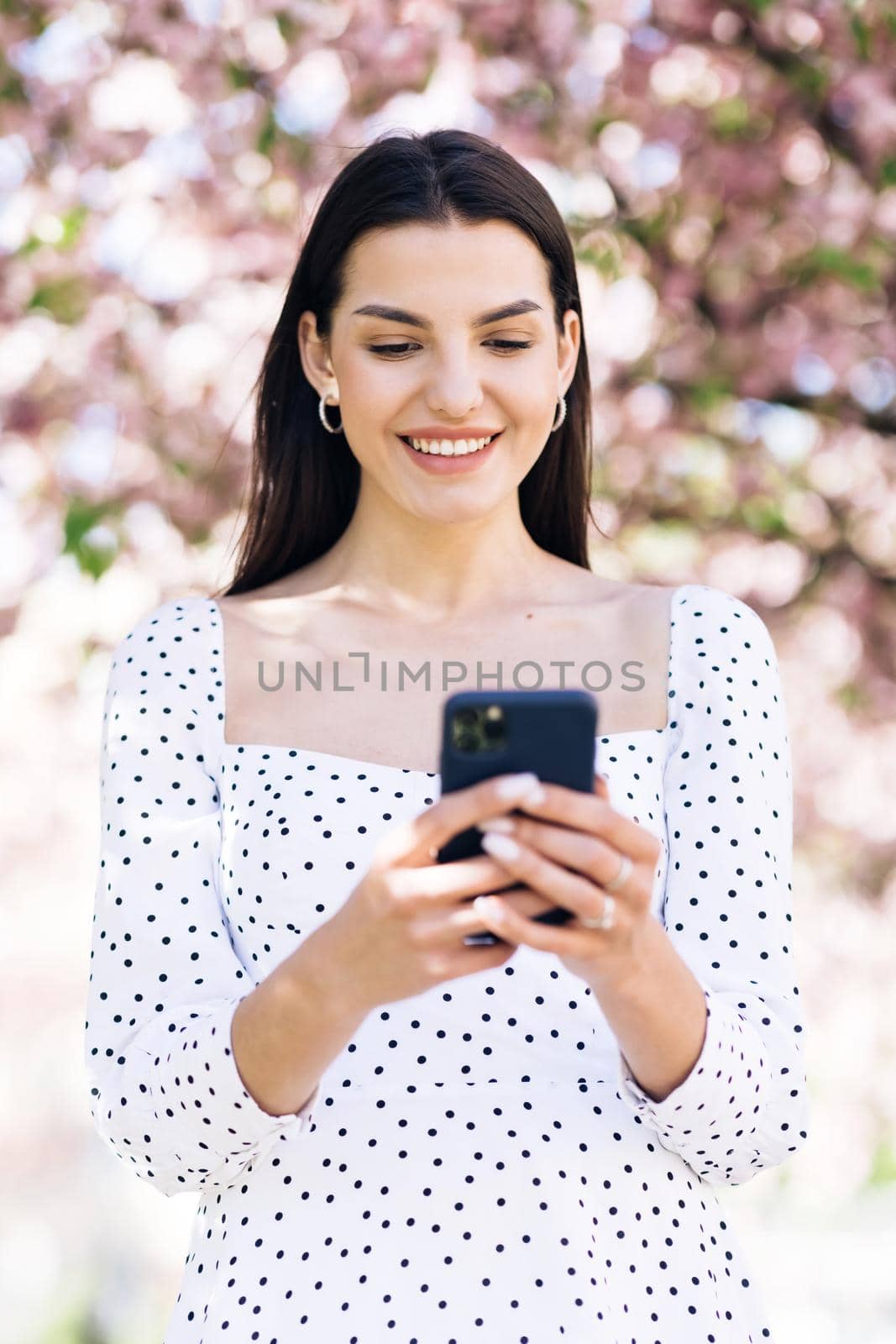 Happy hipster woman typing by mobile phone outdoors. Cheerful girl with smartphone in park on a background of sakura trees. Smiling lady holding cellphone in hands outside.