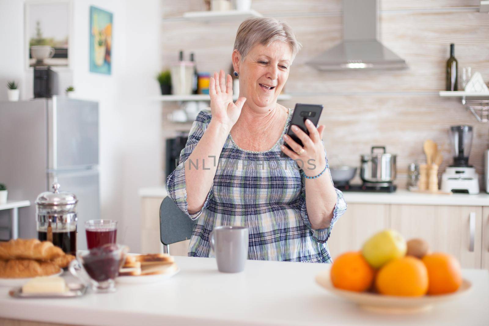 Senior woman waving during video conference using smartphone in kitchen while having breaksfat. Elderly person using internet online chat technology video webcam making a video call connection camera communication conference call