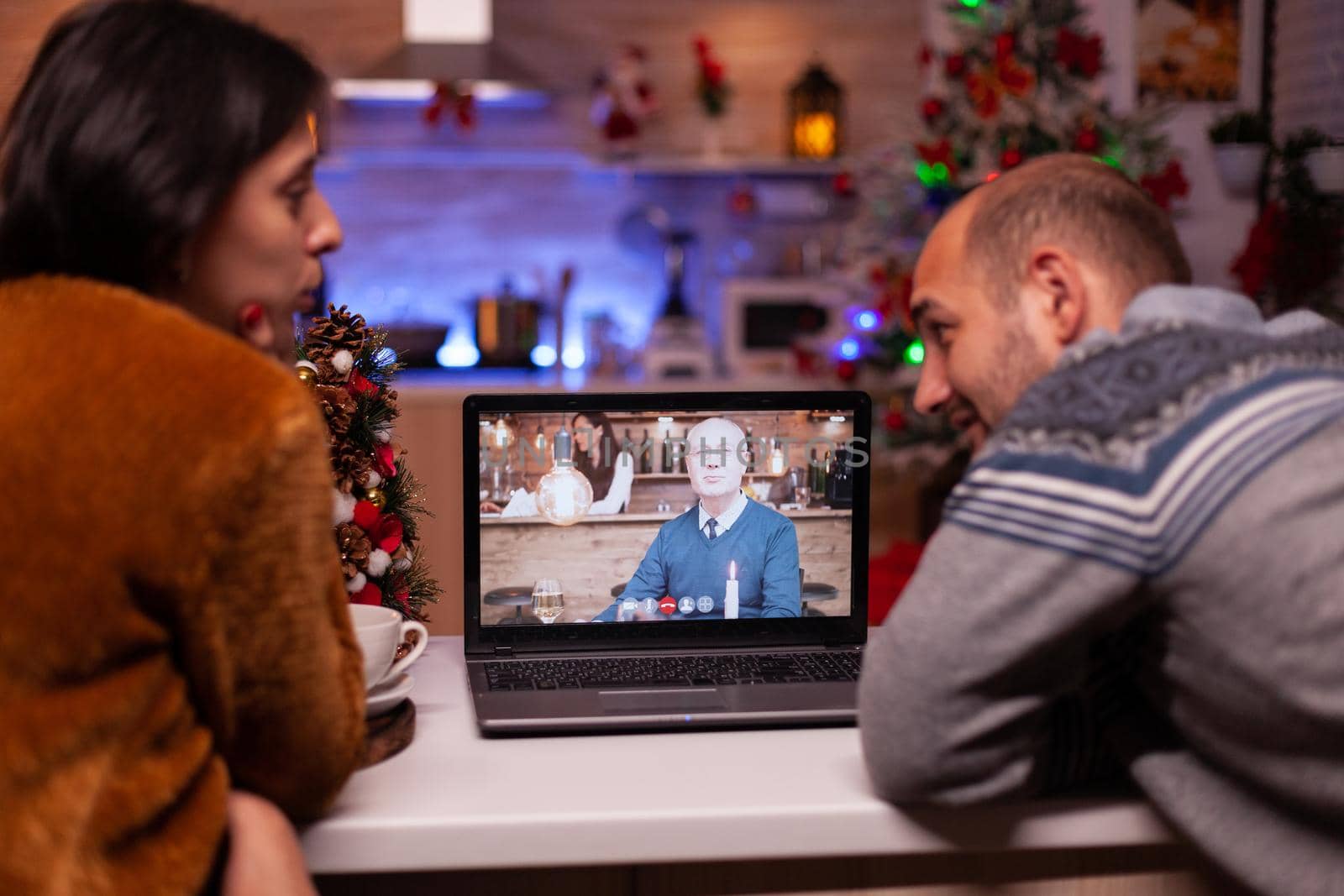 Happy family talking with remote grandfather during online videocall meeting teleconference sitting at table in xmas decorated kitchen. Joyful couple celebrating christmas holiday