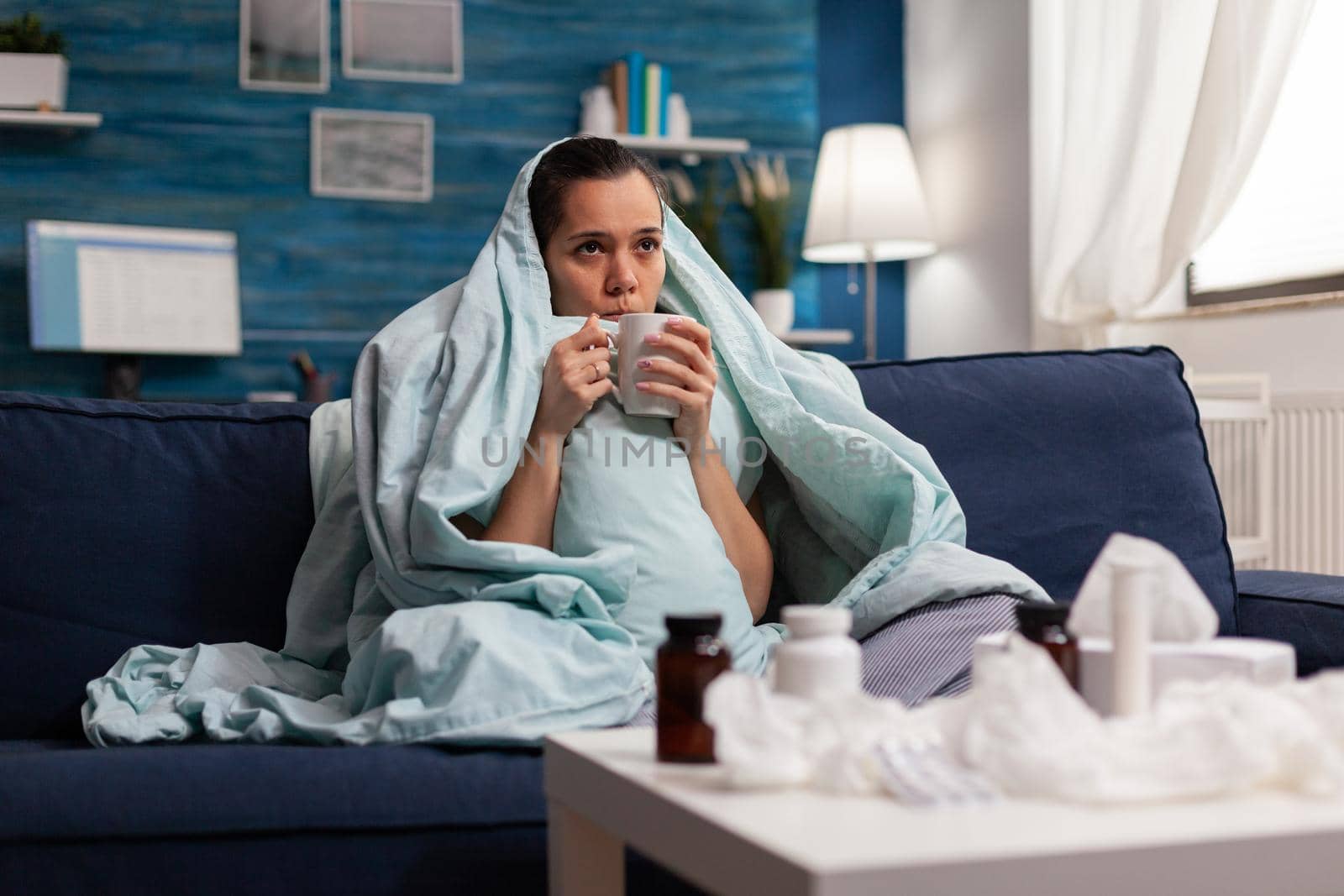 Woman feeling sick in blanket with warm drink at home having seasonal virus symptoms. Unwell young adult in pain headache resting on couch with medical treatment against coronavirus